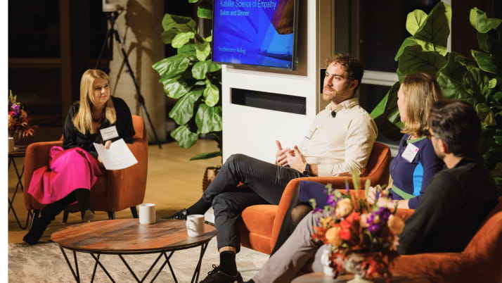 A woman with shoulder-length blond hair wearing a bright magenta skirt sitting in a chair, next to three other people sitting in chairs.