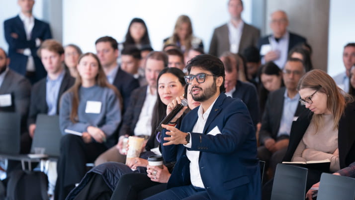 A man with a dark beard and hair among a crowd, gesturing with one hand as he holds a mic in the other.