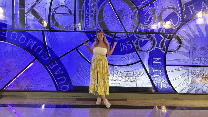 A woman standing against a purple background wearing a white top and long skirt