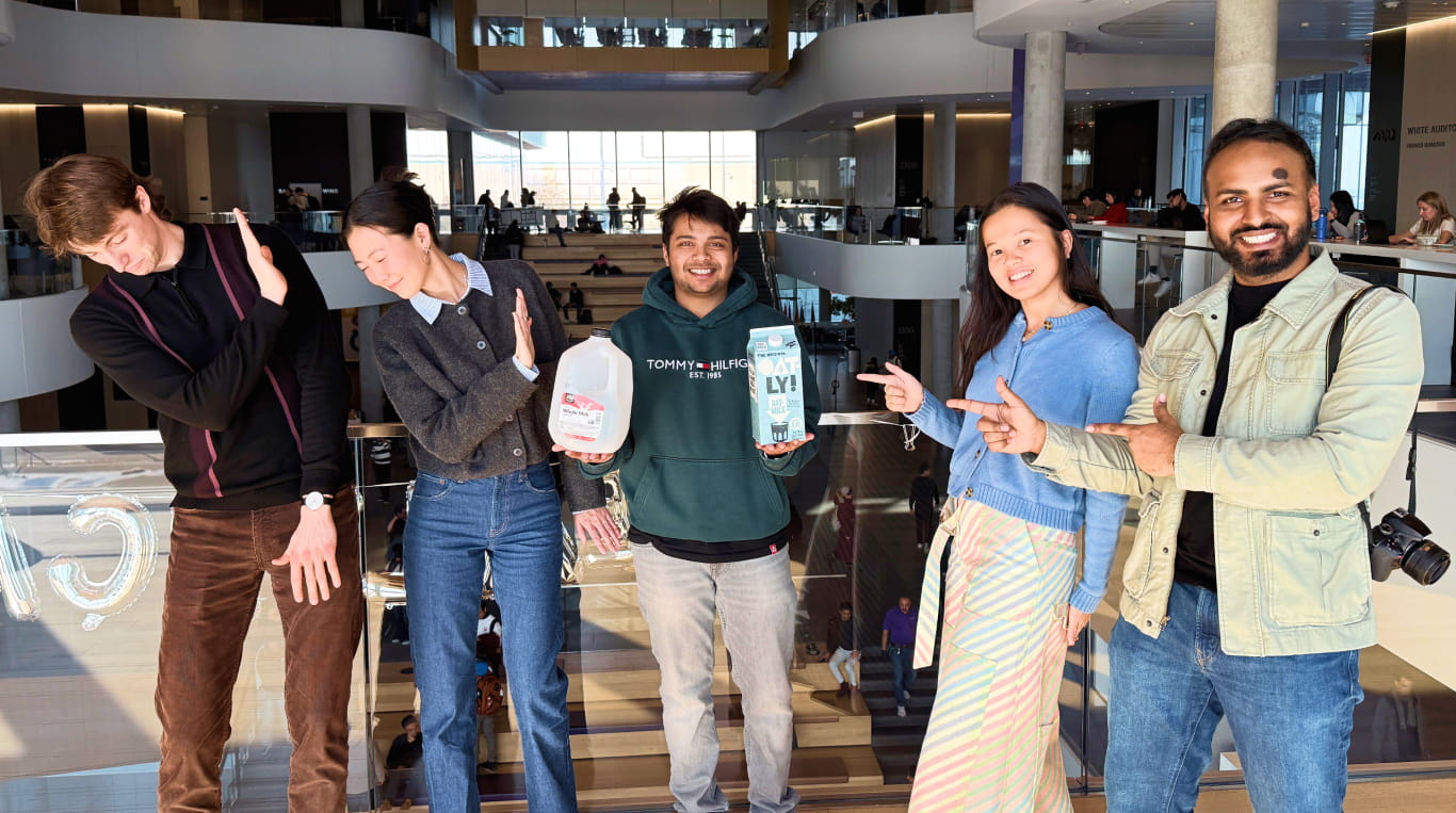 A man stands in the middle of two pairs of people as he holds an empty plastic jug of milk in one hand and a carton of oat milk in the other.