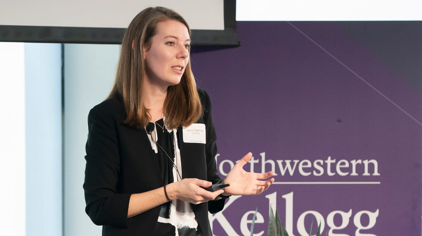 A woman with shoulder-length blonde hair wearing a black blazer and a cream-colored blouse articulating with her hands. 