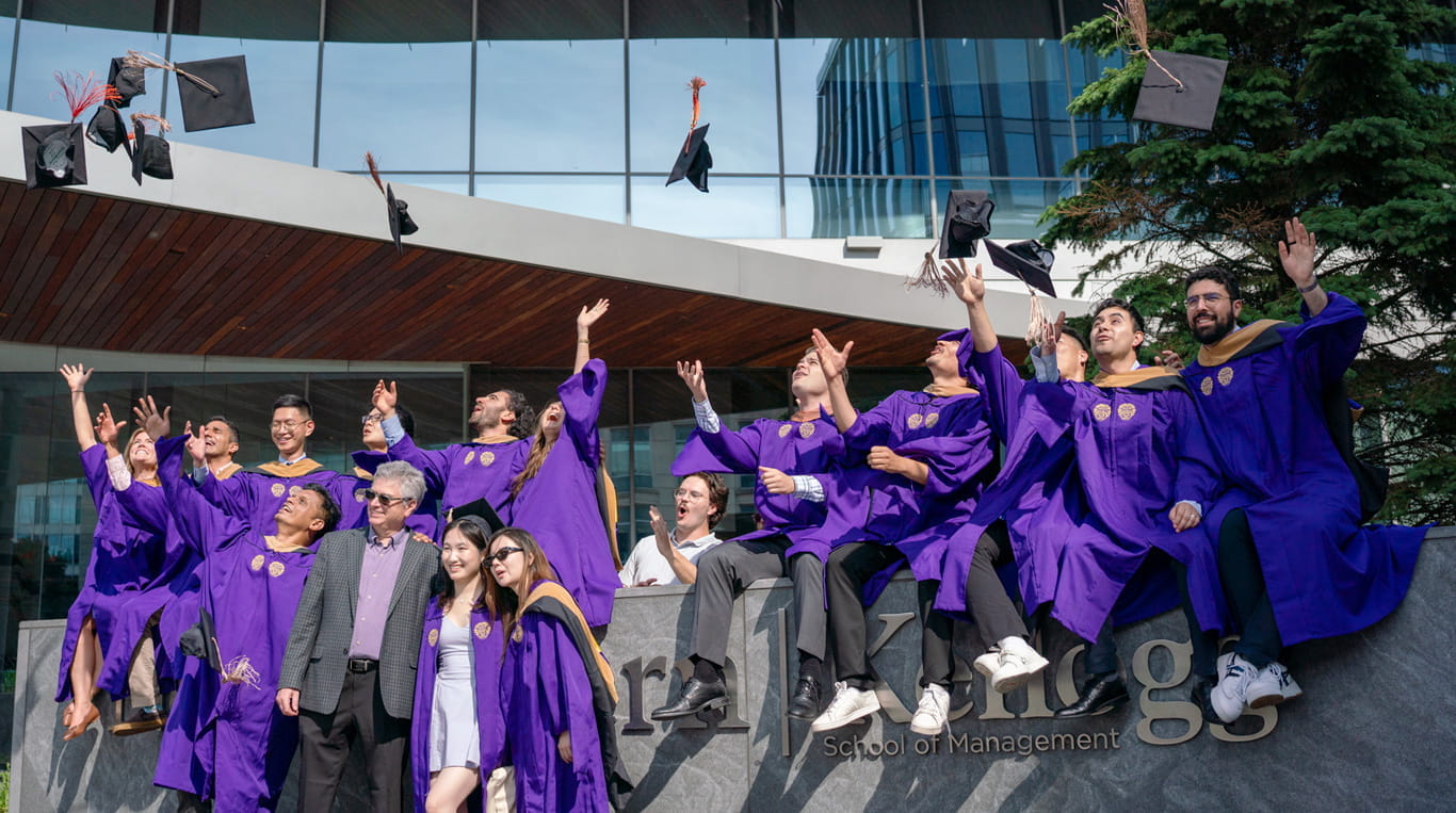 A group of MBA graduates sitting on a ledge and throwing their hats in the air