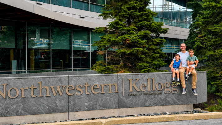 A man and two children sitting atop a sign that reads "Kellogg" in front of a building