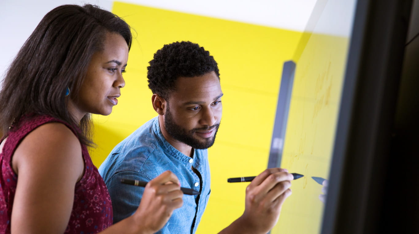 A dark-skinned man writes on a whiteboard, as a dark-skinned woman standing next to him watches.